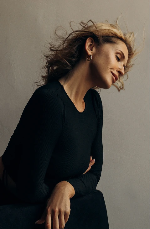 A women wearing all black, leaning over her knee. She has blonde curly hair and hoop earrings on. She has her eyes closed and her head slightly tilted to the side and down. - Facelift in Santa Barbara, CA