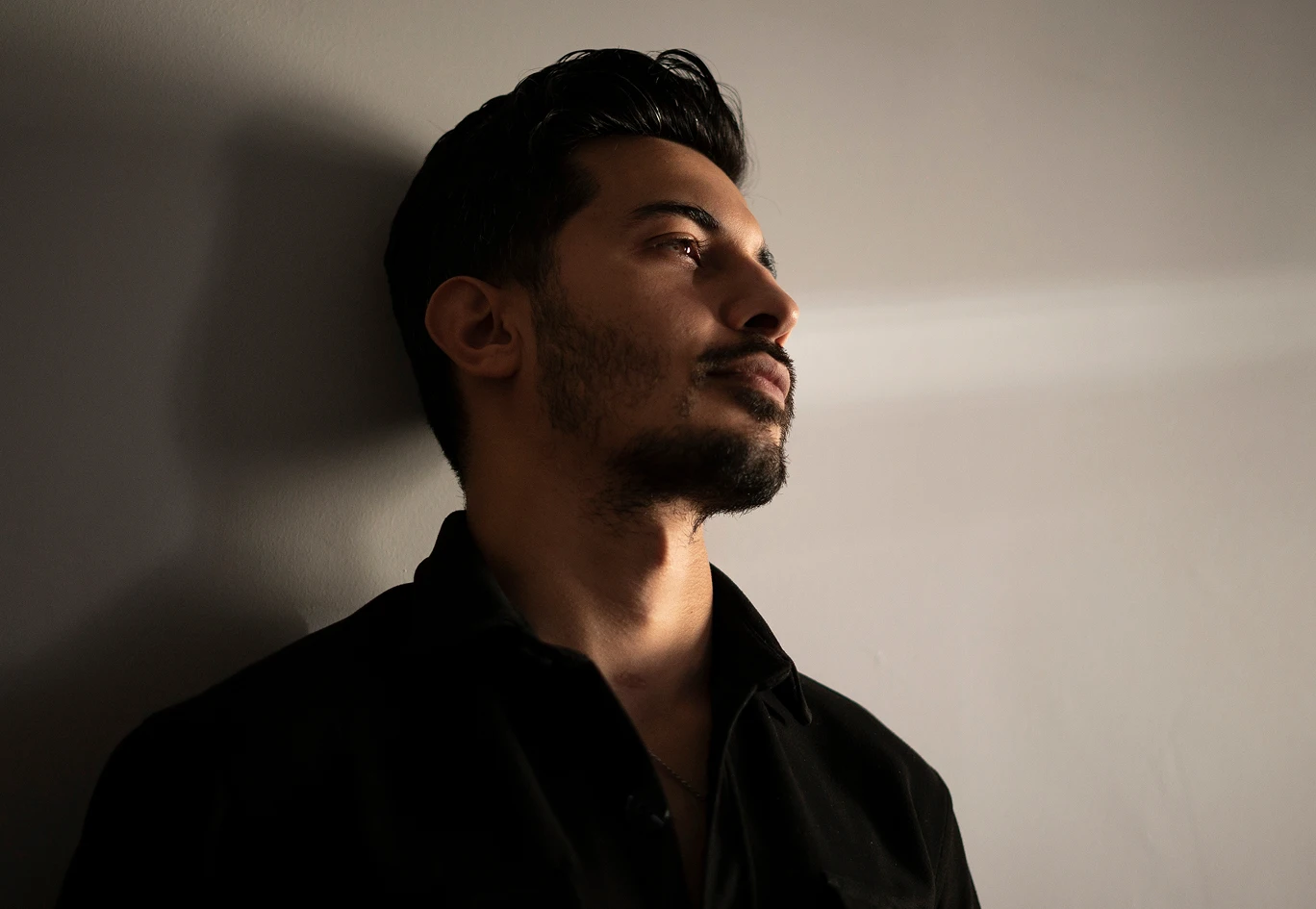 A man is leaning against a wall and wearing a black button up shirt. He looks to the right of the image. He has black hair, a beard and mustache. - Hair in Santa Barbara, CA