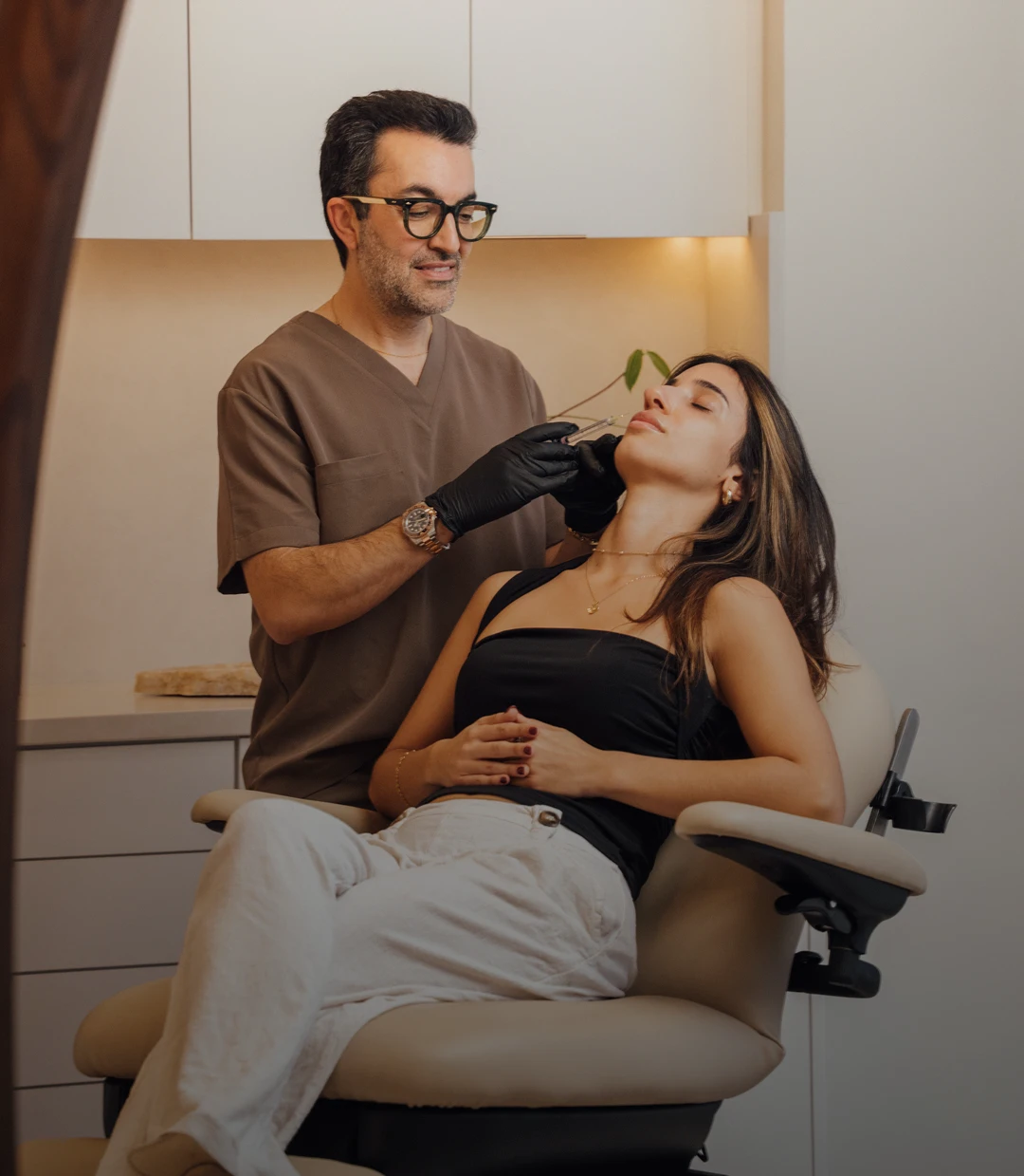 Dr. Moradzadeh about to inject into the patient. The doctor is wearing a brown smock with a gold watch and his glasses. The patient lays in the examination chair in light grey sweats and a black tank top. She has dark brown hair with light highlights, all reflected in a wooden mirror. - Injections in Santa Barbara, CA