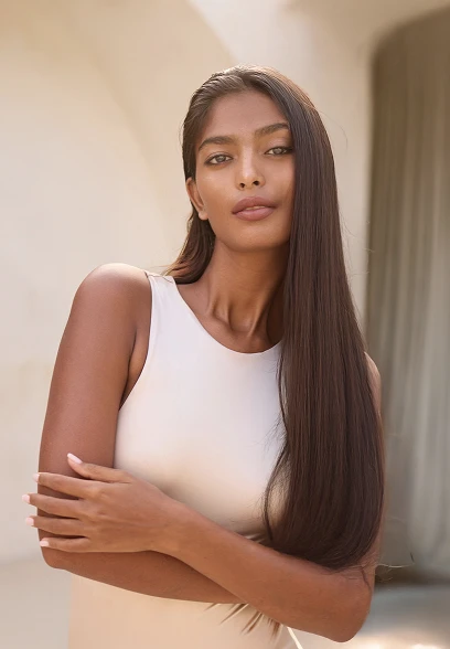 A women in the center of the photo with her arms crossed. She is wearing a white tank top and has long straight dark brown hair. - Liquid Facelift in Santa Barbara, CA