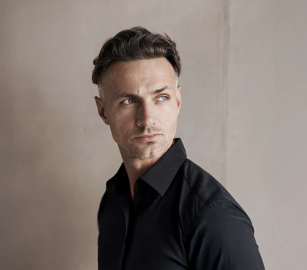 A close up of a man with light eyes and a stubble. He is wearing a black button up shirt. He is looking to the right of the image and has brown hair with greys. - Male facelift in Santa Barbara, CA