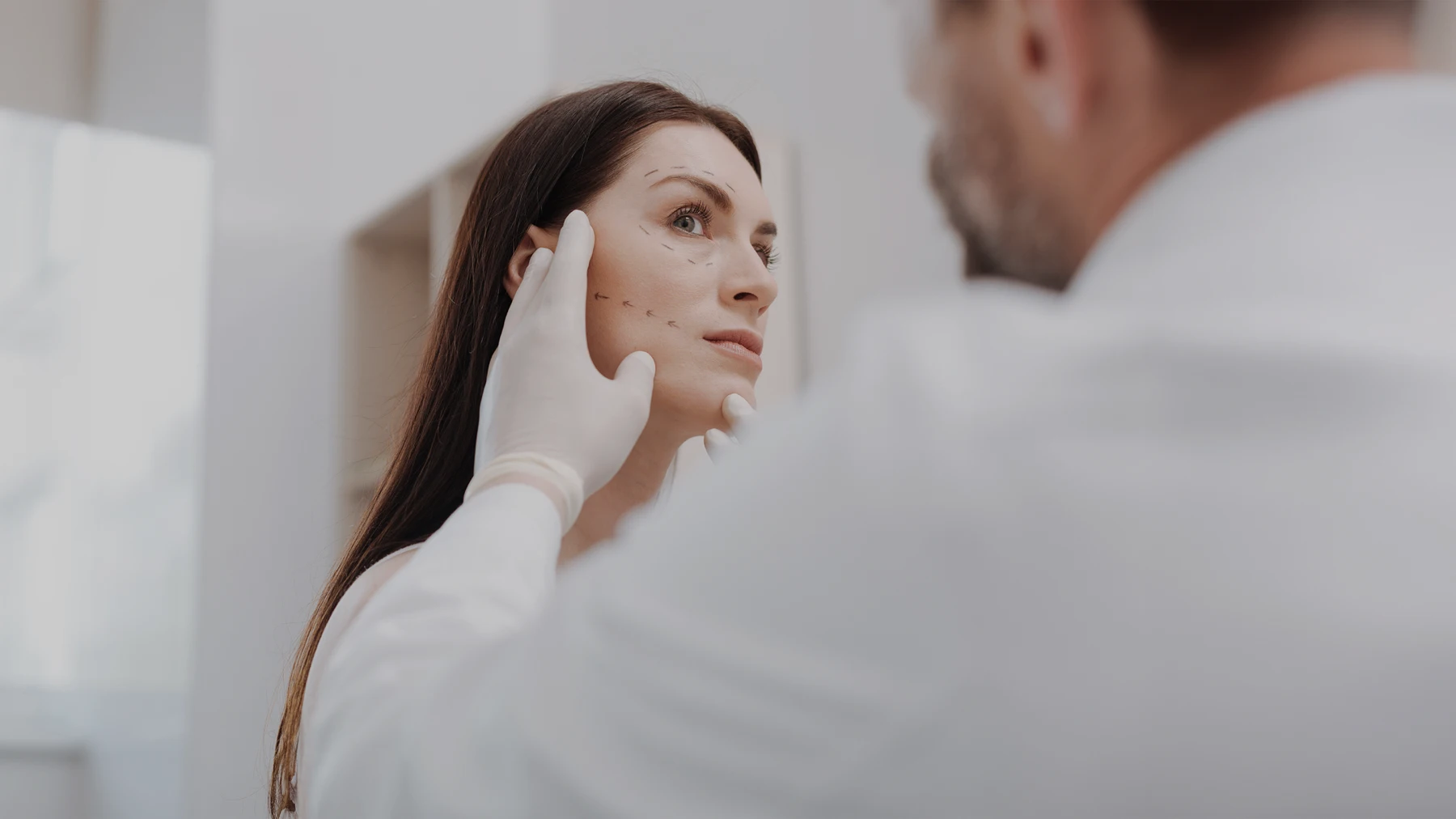 A doctor wearing a white smock and white gloves examines a patient. He has one hand on the patients face. The patient has straight brown hair and marks on her jaw and eyes. - Recon in Santa Barbara, CA