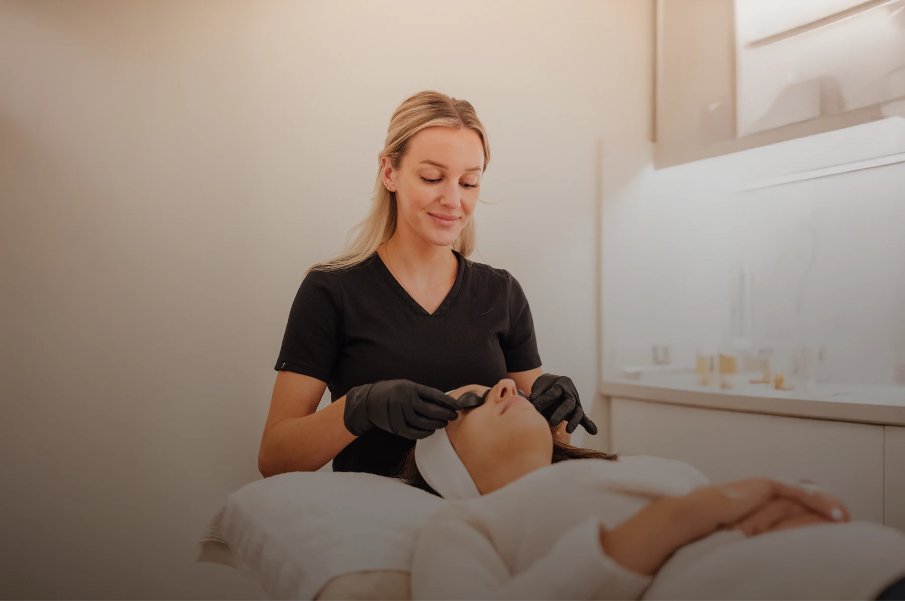 A wide image with a women in the center. The women has blonde hair and looks down on her patients. She is wearing a black smock and gloves. The patient lays down with a white headband on as the women puts tanning goggles on her. - Skin in Santa Barbara, CA