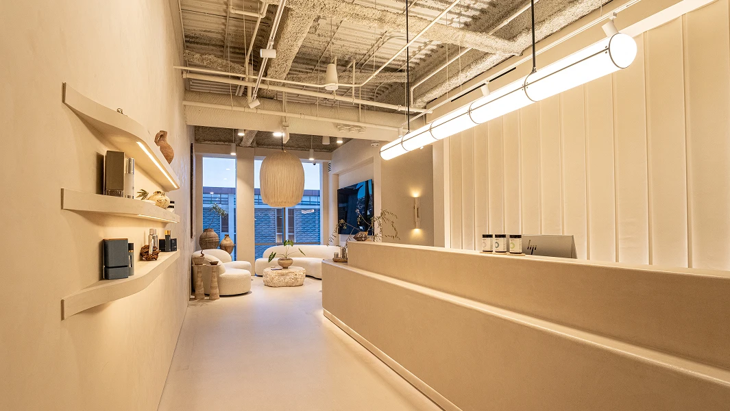 A wide shot of a modern, minimalist medical spa reception area with warm lighting. It features a long, curved limestone desk, floating wooden shelves with skincare products, and a cozy lounge area in the background.