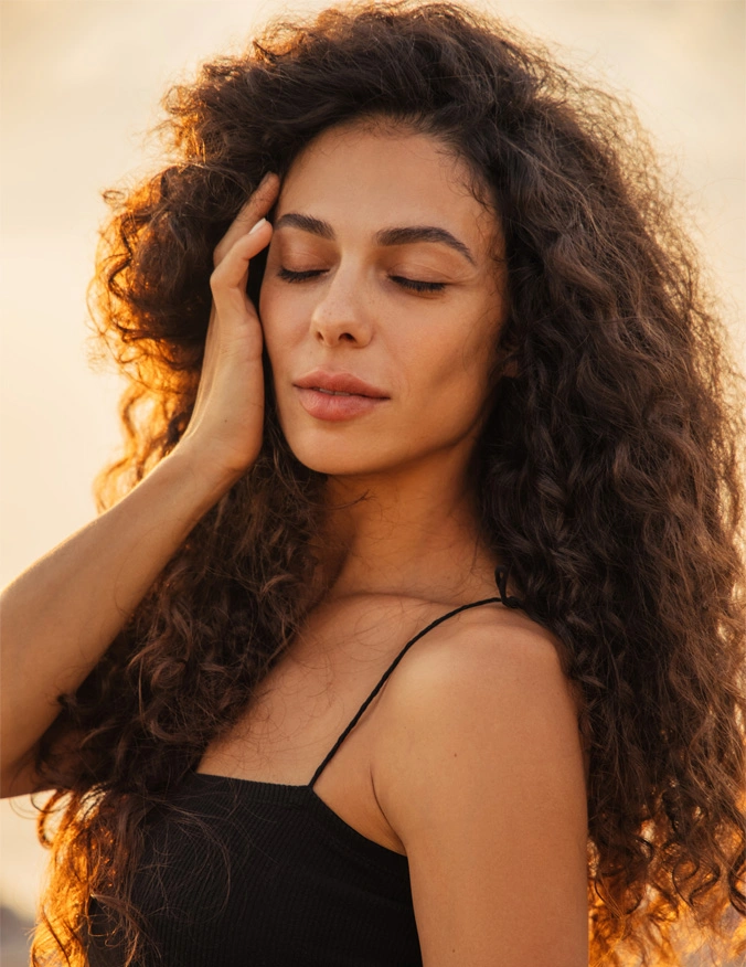 A close up of a women wearing a black tank top. She has long brown curly hair and is touching the side of her face with her eyes closed. - Excel V and Vascular Laser Treatment in Santa Barbara, CA