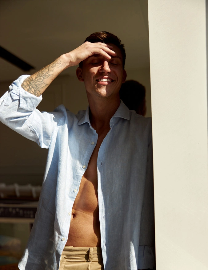 A man appears from a opening and has a light blue button up shirt that is open. He is wearing khakis and is smiling with his eyes closed. He has a hand covering the sun from his eyes and has a clean face with brown short hair. - Hollywood Spectra in Santa Barbara, CA