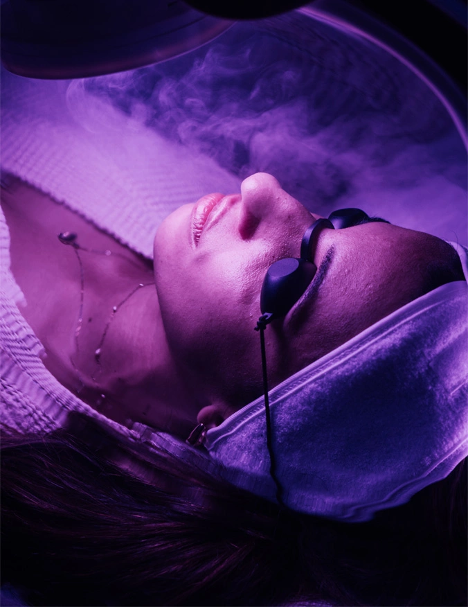 a women in a light therapy bed. The women lays down towards the center of the image, the image is cropped to focus on the lady's face. The had black tanning goggles on and a white headband on. There is a purple light that radiates over the surfaces. - IPL Photo Facial Treatment in Santa Barbara, CA