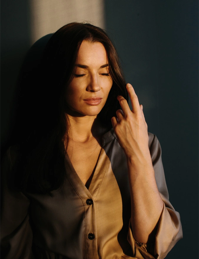 A women stands with a bean of vertical light hitting her. Her eyes are closed and she is wearing a brown button up shirt. She has long dark brown hair and is brushing through it with her left hand. - Kerafactor in Santa Barbara, CA