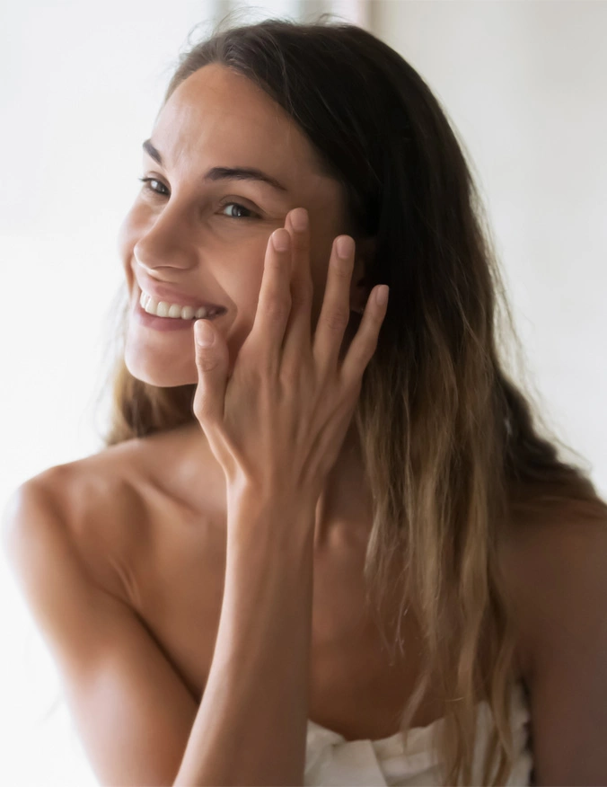 A women looks to the left of the image and smiles. She wears a white bandeau and has long dark hair with blonde highlights. She touches by her eye with her hand. - Lase MD Ultra in Santa Barbara, CA