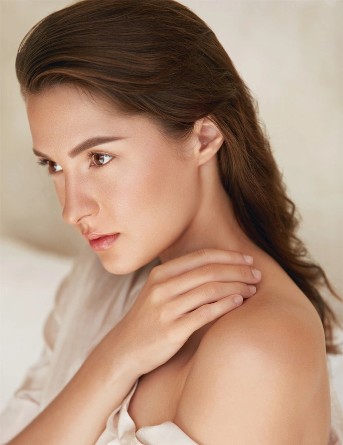 A women touches her shoulder blade and wears a white blouse off one shoulder. She has brown hair and eyes and looks to the left of the image. - Renuvion in Santa Barbara, CA