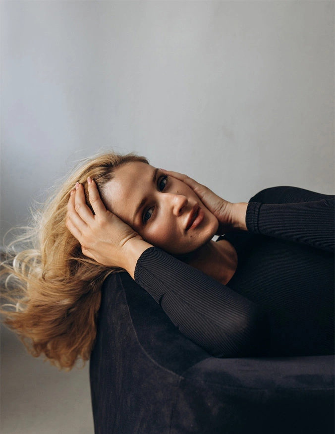 A women holds her head with both hands and is laying on a black chair, she is wearing a black long sleeve shirt and looks directly at camera. She has blonde hair that flows behind the chair. - Brow Lift in Santa Barbara, CA