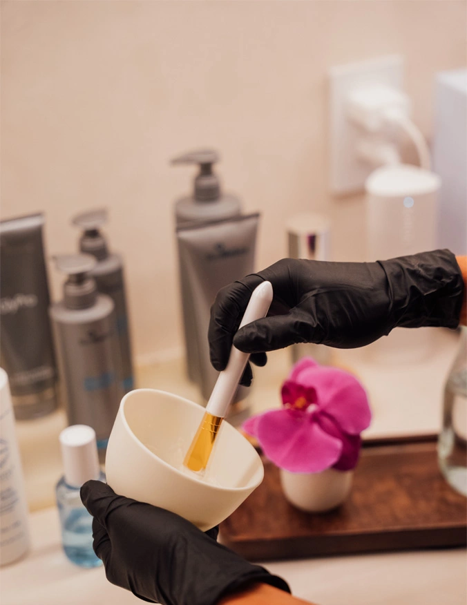 photo featuring multiple products on a counter. The objects in the background are slightly blurred. In this background, we can see a tray with a pink flower on it and a jug with water in it. The foreground shows a set of hands mixing a solution in a white bowl. The hands where black gloves and a white small spatula. - Chemical peels in Santa Barbara, CA.