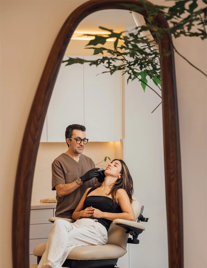 Dr. Moradzadeh about to inject into the patient. The doctor is wearing a brown smock with a gold watch and his glasses. The patient lays in the examination chair in light grey sweats and a black tank top. She has dark brown hair with light highlights, all reflected in a wooden mirror. - Dermal Fillers in Santa Barbara, CA