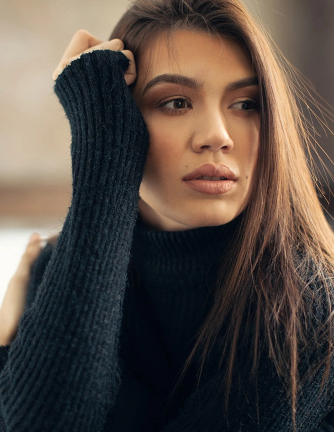 A women wearing a long sleeve turtleneck that is black. She has her hand rested near her eye and has brown straight hair. - Eyelid Surgery in Santa Barbara, CA