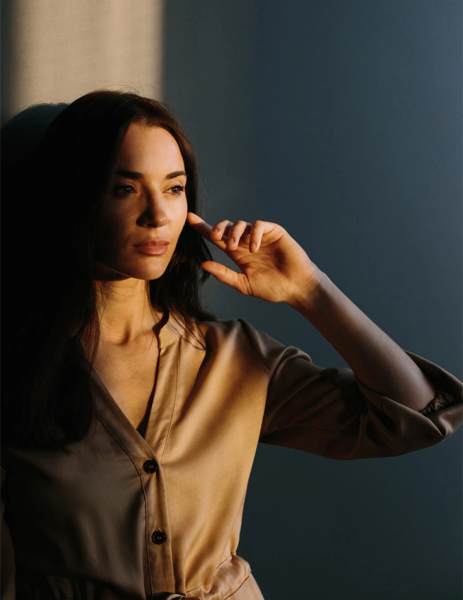 A women stands with a bean of vertical light hitting her. Her eyes are closed and she is wearing a brown button up shirt. She has long dark brown hair and is brushing through it with her left hand. - Facial Trauma Treatment in Santa Barbara, CA