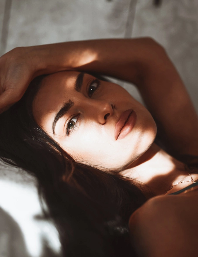a close up of a women's face who is laying with her arm back over her head. She is looking directly into the camera. She has dark brown hair and light eyes. - Functional Rhinoplasty in Santa Barbara, CA