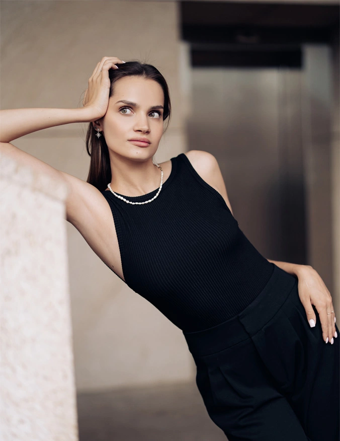 A women leans to the left of the image against a wall. She is wearing a black dress with a small pearl necklace and earrings. She has one hand on her hip and one on her head. She looks off to the left of the image and has dark straight brown hair. - Ellacore 2.0 in Santa Barbara, CA