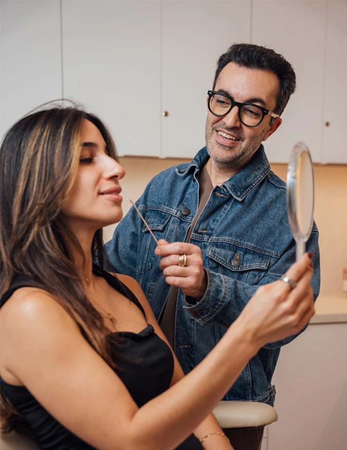 Dr. Moradzadeh is wearing a jean jacket and his glasses. he is holding a long q-tip along the patients nose. The patient has a smile on her face while looking at a mirror she holds in front of her. She has dark brown hair and dark eyes. - Lip lift in Santa Barbara, CA
