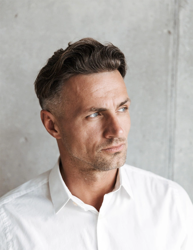A man wearing a white button down shirt in the center of the image. He looks off to the right of the image and has some stubble on his face. He has short brown hair and light blue eyes. - Male Facelift in Santa Barbara, CA