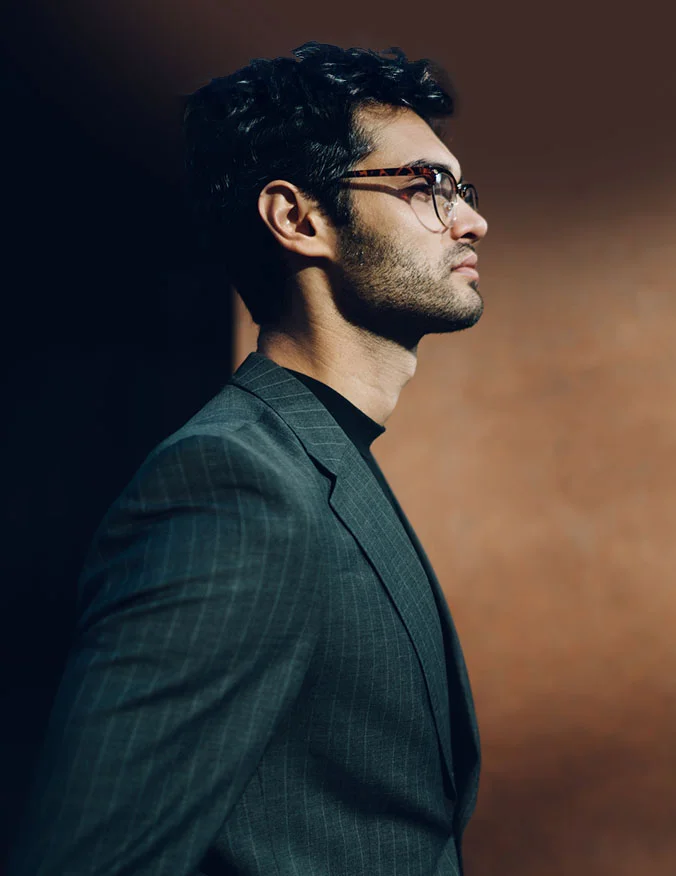 A image of a man looking to the right of the image. The man has black hair and a thin beard and mustache. He is wearing glasses, a black mock neck shirt, and a deep green suit jacket. The background is a brown of fading deepness. - Male Rhinoplasty in Santa Barbara, CA