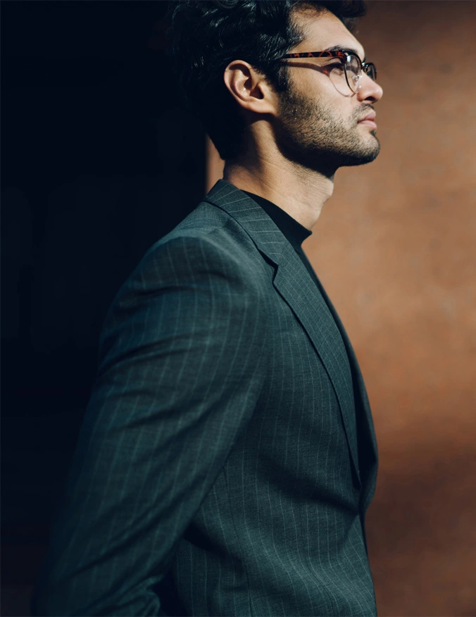 A image of a man looking to the right of the image. The man has black hair and a thin beard and mustache. He is wearing glasses, a black mock neck shirt, and a deep green suit jacket. The background is a brown of fading deepness. - Male Rhinoplasty in Santa Barbara, CA