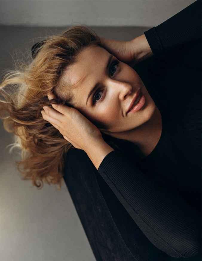 A woman with light brown, tousled hair is viewed from an overhead, slightly angled perspective. She holds her hair with both hands, her head tilted back slightly, and her expression relaxed. She has light skin and light blue eyes. She wears a fitted black top, and the background is a muted gray surface with a soft-focus effect. - Migrane Treatment in Santa Barbara. CA