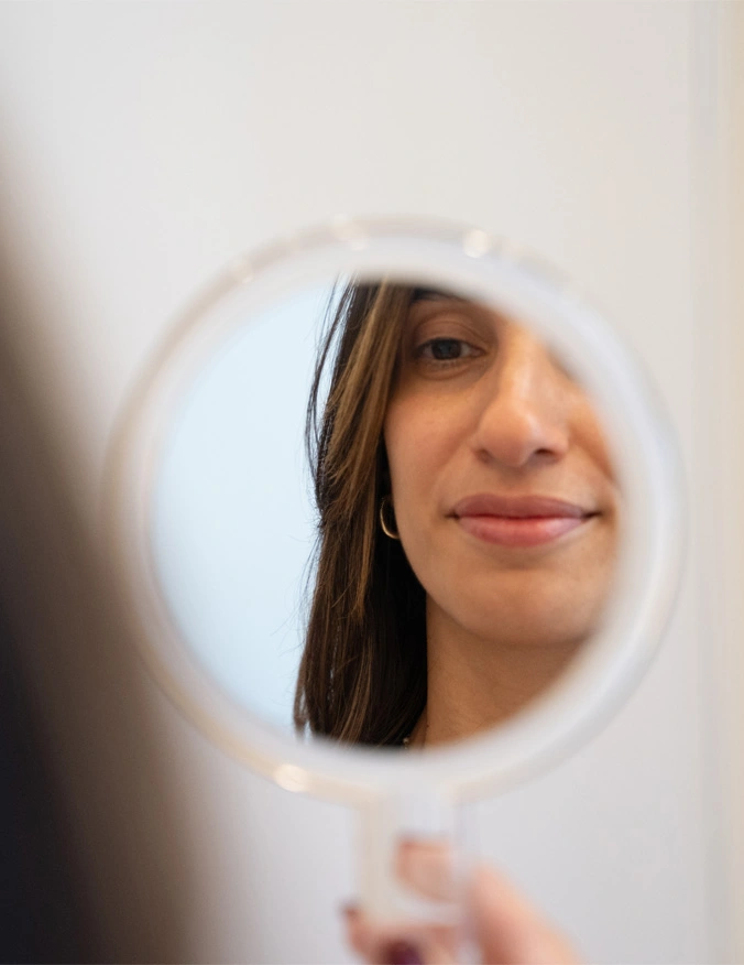 A image with a small circle handheld mirror in the center. In the reflection you can see a women's face with brown hair and a slight smile. - Neck Lift in Santa Barbara, CA