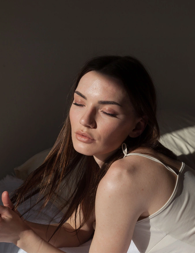 A women laying on a bed and playing with her straight brown hair. She is wearing a white tank top and has her eyes closed. - Non surgical rhinoplasty in Santa Barbara, CA