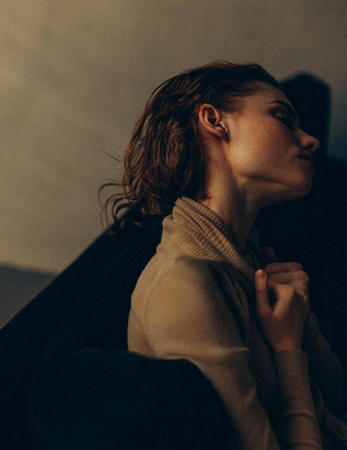A women wearing a cream turtleneck and lays back on a black couch. She leans her head to the side and grips at the collar of her shirt. She has short brown curly hair. - Otoplasty in Santa Barbara, CA