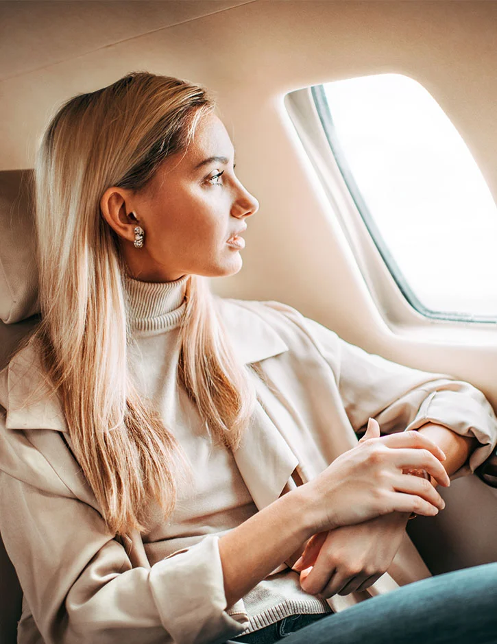 A blonde women with dark roots and eyebrows looks out a airplane window. She has sparkly silver earrings on and her mouth is slightly open. She is wearing a cream sweater with a tan jacket over top. Her left elbow is on the arm rest while her right hand softly touches her left wrist. - Out of Town in Santa Barbara, CA.