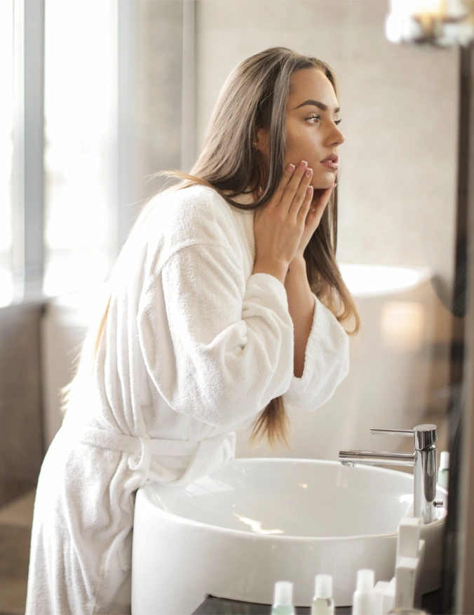 A women leans over a white sink and looks into a mirror. She is in a robe and touching both sides of her face. She has long brown hair. - PRP with microneedling in Santa Barbara, CA