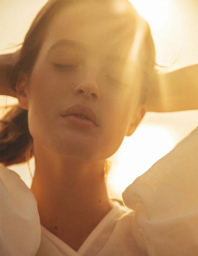 A strong sun beam comes from the top of the image. The image is a close up of a women wearing a white blouse. Her eyes are closed and her brown hair pulled back by both of her hands. - Red light therapy in Santa Barbara, CA