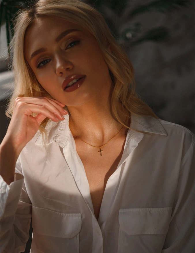 A women with a glow of light in the left middle of the image. She is wearing a white button up shirt and has a gold cross necklace. She has blonde curly hair and looks into the camera. - Revision Rhinoplasty in Santa Barbara, CA