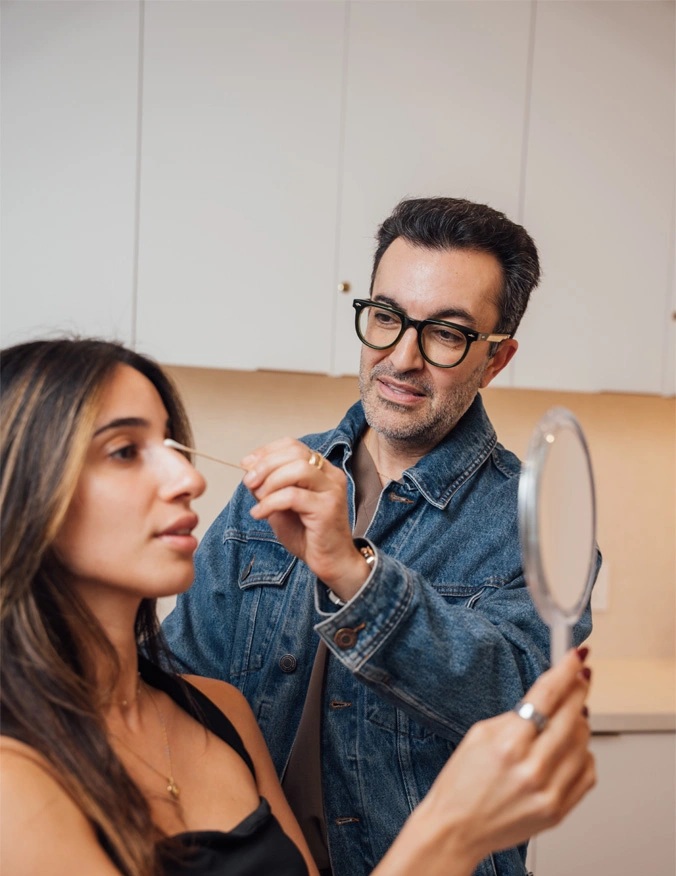 Dr. Moradzadeh is wearing a jean jacket and his glasses. he is holding a long q-tip along the patients nose. The patient has a smile on her face while looking at a mirror she holds in front of her. She has dark brown hair and dark eyes.