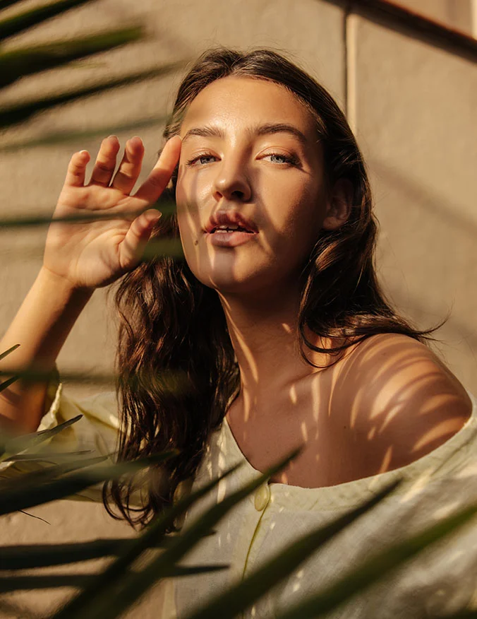 A image of a women with her hand raised by her eyebrow. She has brown hair with slight curls and looks directly into the camera. She is wearing a white blouse with one side off her shoulder. - Septoplasty in Santa Barbara, CA