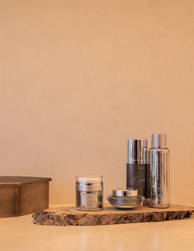 Four products sit on a small marble slab. The products vary in size, and on the side of the products is a small chest. The background is a cream tan color. - Skin care in Santa Barbara, CA