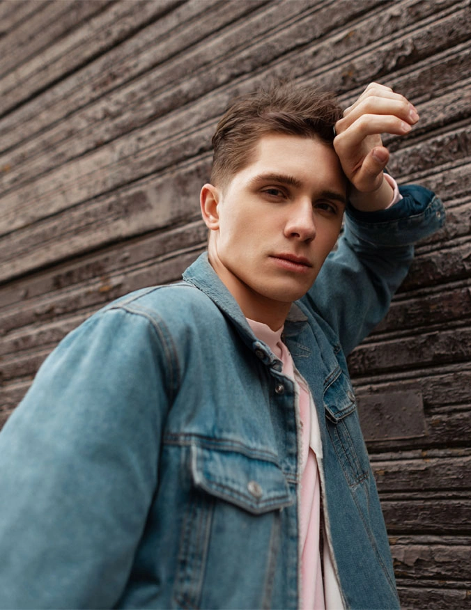 A image with a man leaning against a wood wall. He is wearing a light pink shirt and a jean jacket over top. He has a clean face and short brown hair. - Teenage Rhinoplasty in Santa Barbara, CA